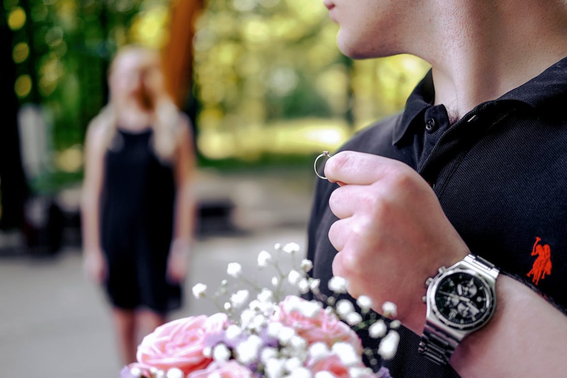 Soft focus of young male in casual clothing holding wedding ring and bouquet of flowers while going to make proposal to woman in garden on blurred background