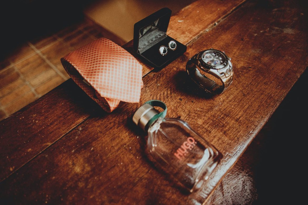 From above composition of silk tie above male perfume bottle and steel watch with cufflinks located on dark wooden surface